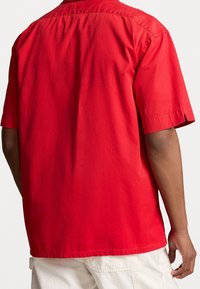 Red short-sleeve shirt with a relaxed fit, made of lightweight fabric. Features a straight hem and small side slits at the sleeves.