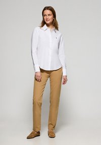 Camicia bianca con colletto e logo, abbinata a pantaloni beige e mocassini marroni. Il tessuto appare liscio con una vestibilità sartoriale.