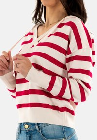 Hoodie rayé rouge et blanc avec un design en V, texture tricotée et larges poignets. Présente un cordon de serrage au niveau du cou et un accent de logo.