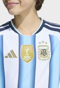Osoba koja nosi argentinsku plavu i bijelu prugastu nogometnu majicu s zlatnom oznakom FIFA Svjetski prvak 2022 i Adidas logom na prsima.
