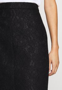 Close-up van de taille en hand van een persoon die een hooggetailleerde zwarte rok met bloemrijke kanttextuur en een wit topje draagt.