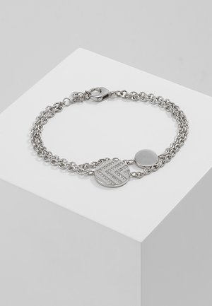 Zilveren dubbele kettingarmband met twee ronde bedels, waarvan één met een chevronpatroon en kleine kristallen, tentoongesteld op een witte ondergrond.