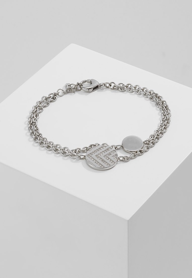 Bracelet double chaîne en argent avec deux breloques rondes, dont une ornée d'un motif chevron avec de petits cristaux, présenté sur une surface blanche.