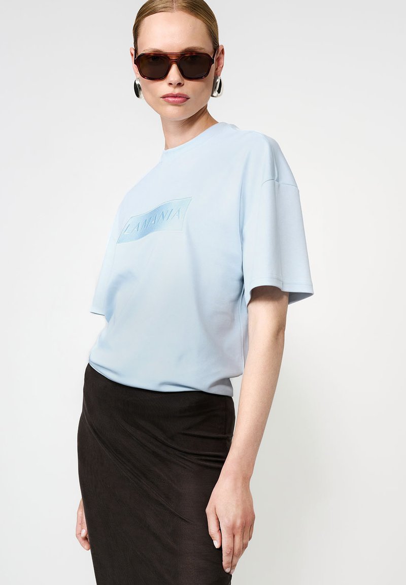 Moteris, vilkinti šviesiai mėlyną LA MANIA marškinėlį, juodą sijoną, dideli sidabriniai auskarai ir tamsūs per dideli saulės akiniai, pozuoja prieš baltą foną.