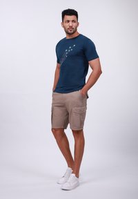 Camiseta de algodón azul oscuro con diseño de avión, combinada con pantalones cortos beiges de cargo con múltiples bolsillos y zapatillas blancas.