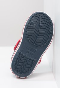 Zoccoli rossi con una suola in gomma blu texturizzata, caratterizzata da profonde scanalature per una buona aderenza, un bordo bianco e un logo del marchio prominente sulla parte inferiore.