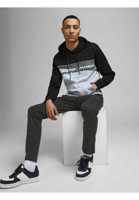 Zwarte en grijze hoodie met logo, gecombineerd met donkergrijze broek. Model draagt marineblauwe sneakers met witte accenten. Eenvoudige achtergrond.