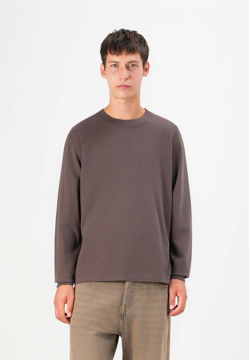 Cămașă lungă, maro, din bumbac moale, cu guler rotund și croială lejeră, asociată cu pantaloni bej deschis. Design minimalist.