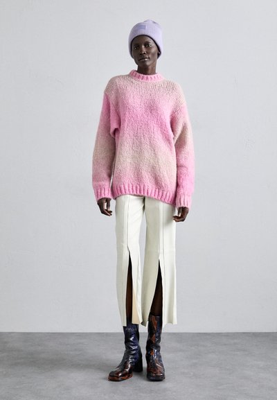Maglione rosa oversize lavorato a maglia con scollo a coste, abbinato a pantaloni crema con spacchi laterali. La modella indossa un berretto lavanda e stivali con fantasia.