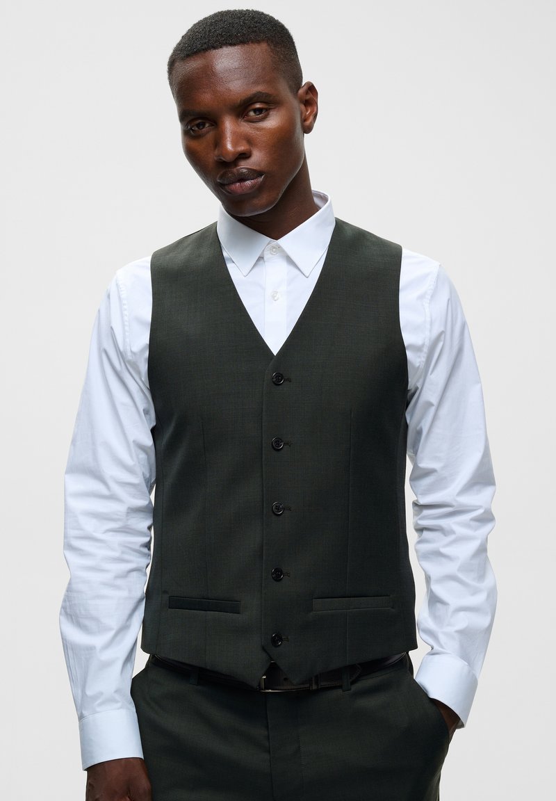 Homme portant un gilet vert foncé boutonné sur une chemise blanche, les mains dans les poches, sur un fond clair uni.