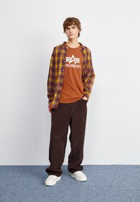 Flanel košulja u ljubičasto-žutom kariranom uzorku, narančasta majica s logom, smeđa pamučna hlače, bijele tenisice, stoji na svijetloplavom tepihu.