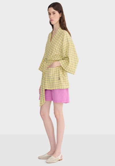 Femme debout les mains dans les poches, portant un haut cache-cœur à carreaux jaunes avec de larges manches, un short rose et des chaussures beige tissées.