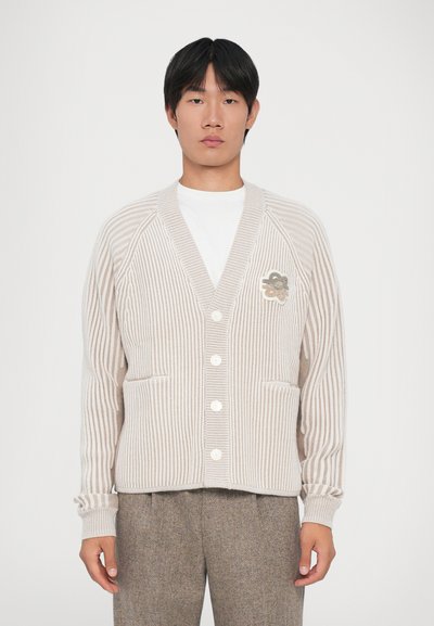 Ung mand iført en beige og hvid stribet cardigan med knapper, en hvid skjorte og brune strukturerede bukser mod en ensfarvet baggrund.