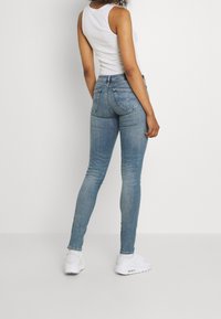 Jeans ajustados de denim azul claro, con cintura de altura media, piernas ceñidas y dos bolsillos traseros. Combinados con una camiseta sin mangas blanca y zapatillas.