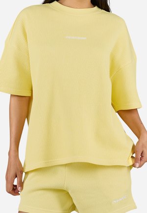 Kobieta ubrana w żółtą, prążkowaną bluzkę z krótkim rękawem i dopasowane spodenki, oba z małymi białymi logo "PEGADOR", pozująca na jednolitym tle.