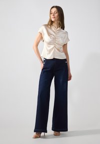 Top corto a maniche corte in seta color crema con parte anteriore arricciata, abbinato a jeans in denim scuro a gamba larga con dettagli in bottoni e cuciture a contrasto.
