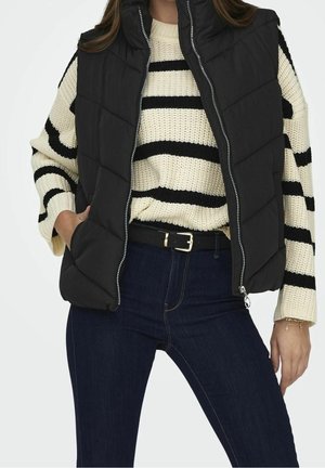 Femme portant une veste matelassée noire sur un pull rayé crème et noir avec un jean bleu foncé et une ceinture noire, main dans la poche de la veste.
