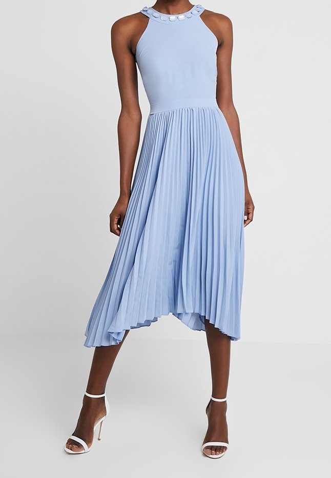 Femme portant une robe midi plissée bleu clair sans manches avec un décolleté rond orné d'embellissements circulaires, associée à des sandales blanches à talons hauts.