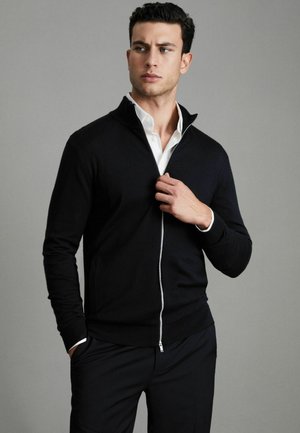 Juoda zip-up megztinis su aukštu apykakle, pagamintas iš švelnios medžiagos. Turi sidabrinę užtrauktuką ir kontrastingą baltą marškinėlį po juo.