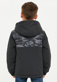 Chlapec v tmavé bundě s maskáčovým vzorem na horní části zad a kapuci, stojící zády k fotoaparátu na jednobarevném pozadí.