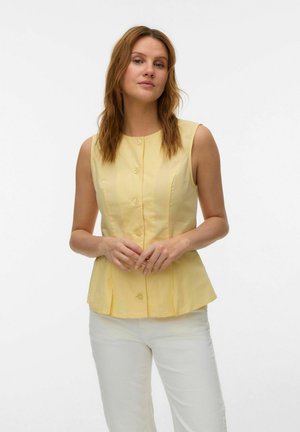 Femme portant un haut jaune sans manches à boutons et un pantalon blanc, debout les mains jointes devant un fond blanc uni.