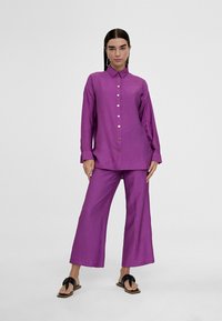 Camisa morada de botones con cuello, combinada con pantalones morados de pierna ancha. La tela tiene un acabado texturizado; las sandalias presentan adornos de lazos.