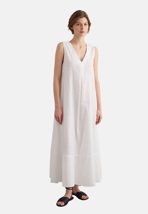 Mujer de pie con un vestido largo blanco sin mangas con escote en V y sandalias planas negras sobre un fondo liso y claro.