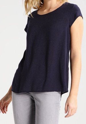 Femme portant un t-shirt bleu marine à manches courtes avec de petits pois blancs et un jean gris clair ajusté, debout devant un fond uni.
