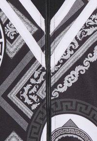 Svart och vitt tyg med utsmyckade barockstilsvirls, grekiska nyckelmönster och en central svart dragkedja som löper vertikalt.