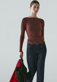 Kvinde iført en tætsiddende brun langærmet top og mørkeblå jeans, der holder en sort taske med et rødt tørklæde mod en ensfarvet baggrund.