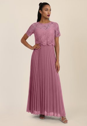 Frau trägt ein mauvefarbenes Spitzenkleid mit kurzen Ärmeln und einem plissierten langen Rock, steht mit einer Hand auf der Hüfte und silbernen High Heels.