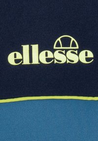 Námořnicky modrá mikina s výrazným žlutým logem „ellesse“, s kontrastní modrou spodní částí a žlutým lemem podél spodního okraje.