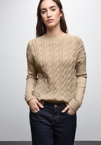 Maglione beige a maglia rasata con scollo rotondo e polsini a coste, abbinato a jeans in denim scuri con chiusura a bottone e tasche laterali.