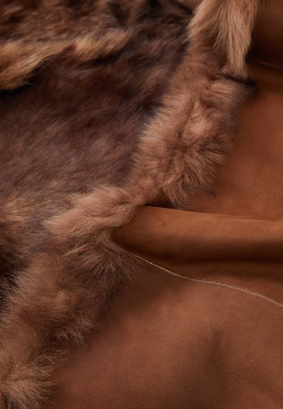 Mehakav rjav shearling material, ki prikazuje puhasto krzno z obloženjem in gladko zamškasto zunanjostjo z vidnimi detajli šivanja.