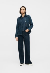 Femme debout, une main dans la poche, portant une chemise et une veste en denim bleu foncé, un jean à jambes larges, et des baskets noires et blanches.