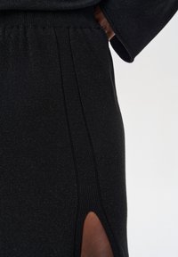 Czarny dzianinowy element garderoby o teksturowanym wykończeniu, z bocznym rozcięciem i ściągaczem w pasie, prezentujący gładki, błyszczący wygląd.