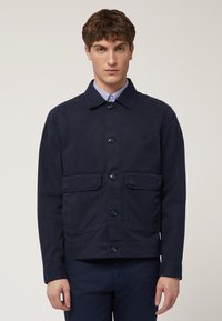 Marineblauwe jas van een gladde, duurzame stof. Heeft een knoopsluiting aan de voorkant, twee grote voorzakken en een klassieke kraag.