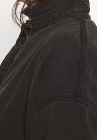 Chaqueta de mezclilla negra con un cuello alto, con cierre de botones y detalles de costura visibles. La tela tiene una apariencia texturizada.
