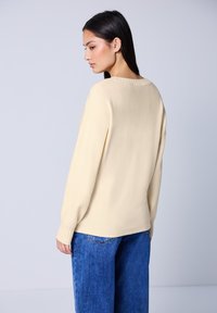 Mujer con cabello largo y oscuro, vistiendo un suéter crema y jeans azules, vista desde la parte de atrás sobre un fondo claro y liso.