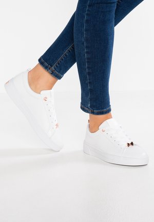 Person wearing dark blue skinny jeans and white low-top sneakers with rose gold accents on a white floor.