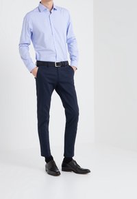 Camisa azul claro de botones con un pequeño patrón de puntos, combinada con unos pantalones ajustados azul marino y unos zapatos de vestir negros. Cinturón negro con hebilla de plata.