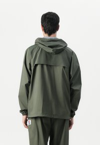Giacca leggera verde oliva con cappuccio, polsini regolabili e una aletta di ventilazione sul retro. Presenta una patch del logo vicino all'orlo.