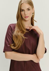 Blondína s dlhými vlasmi, oblečená v krátkoplej mauve top, s perlovými náušnicami, pózujúca s jednou rukou pri tvári na neutrálnom pozadí.