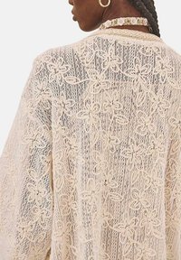 Cárdigan de encaje en color crema con patrones florales, textura de punto abierto, cae holgadamente, con un cuello amplio y hombros caídos.