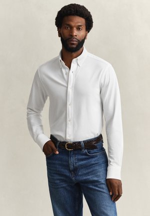 Mann med skjegg iført hvit skjorte med knapper, stukket inn i blå jeans med flettet mørkebrun belte, stående med den ene hånden i lommen.