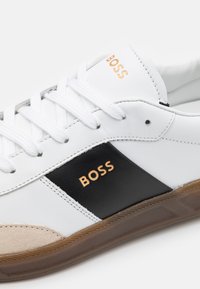 Vita sneakers i läder med svarta detaljer, med en guldfärgad "BOSS"-logotyp, vita skosnören och en texturerad gummisula med brun kant.
