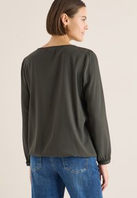 Donna con capelli castani corti che indossa una blusa a maniche lunghe larga di colore verde oliva scuro e jeans blu, in piedi di profilo su uno sfondo chiaro uniforme.