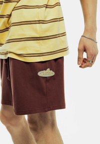 Bruine katoen shorts met een elastische tailleband en een trekkoord, met een lichtgekleurde logopatch op de linker dij. Gestreept geel shirt erboven.