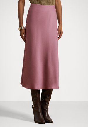 Femme portant une jupe midi en satin mauve associée à des bottes à talons hautes jusqu'aux genoux de couleur brun foncé et des bracelets dorés à un poignet.