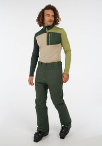 Groen en beige sportieve zip-up top met lange mouwen en een rits borstzak, gecombineerd met donkergroene broek en skibotten.